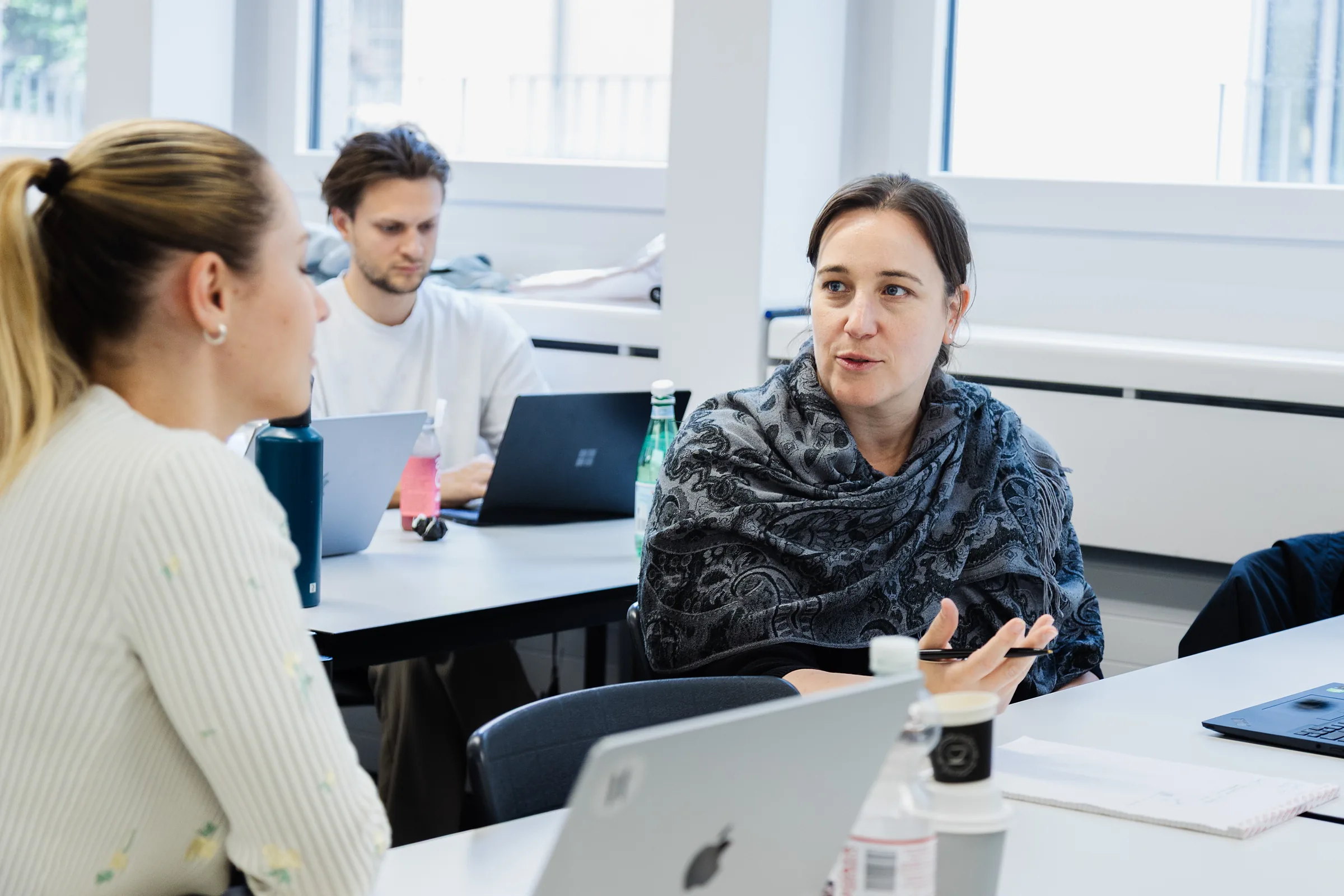 Participant talking with the person next to her in the classroom and gesturing with a pen, two participants working on laptops behind them